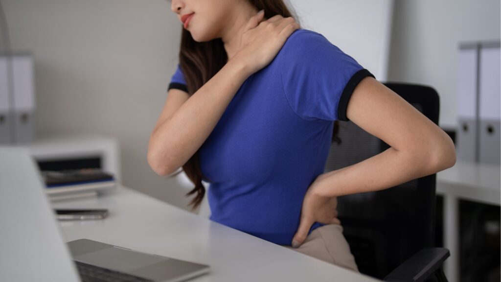 Office worker experiencing neck and lower back pain from prolonged sitting, highlighting workplace ergonomics risks in Singapore offices