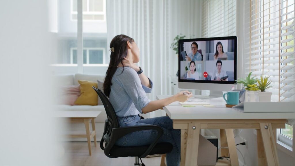 Employee working at a home desk during a video meeting, holding her neck to relieve discomfort from prolonged screen-based work.