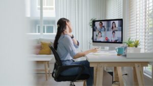Employee working at a home desk during a video meeting, holding her neck to relieve discomfort from prolonged screen-based work.