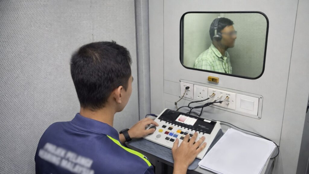 Workplace hearing assessment in progress, with a technician operating audiometric equipment while an employee undergoes testing in a soundproof booth
